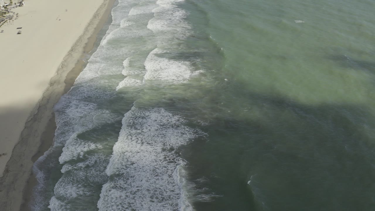 Oblique aerial view of green water and white surf meeting a pale Los Cabos beach, with no visible people in the frame.