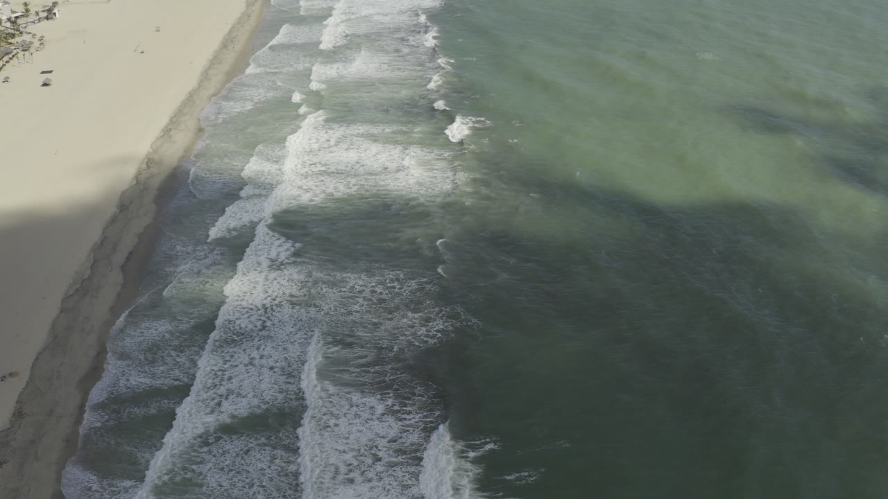 Aerial view over Los Cabos surf, with white waves breaking diagonally along pale sand and small people faintly visible near the top of the beach.