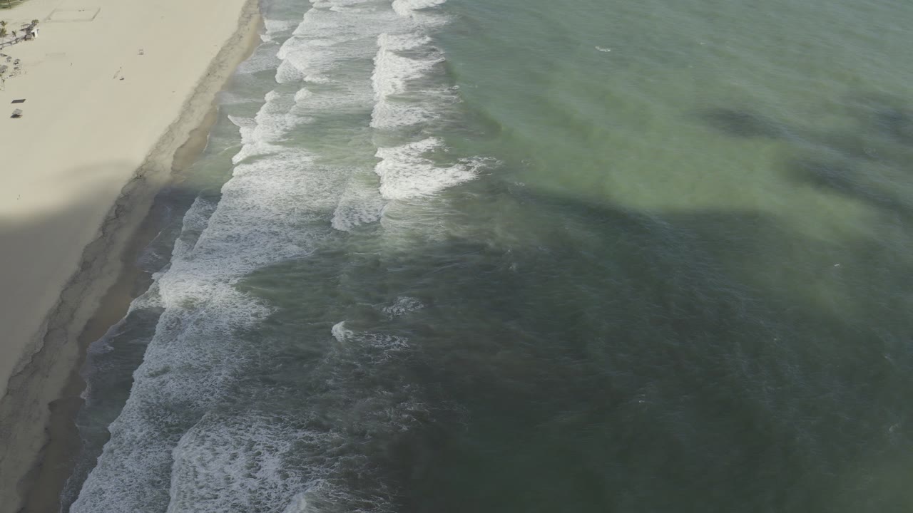 Aerial view over a Los Cabos shoreline with pale sand at left, green-blue water at right, white surf bands near the beach, and no visible people.