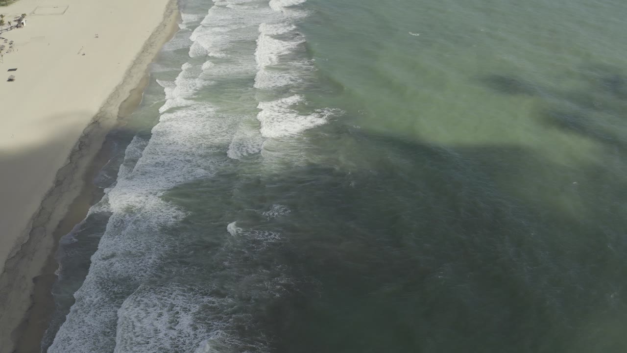High overhead frame of the Los Cabos coast with foamy waves advancing along the sand on the left and darker green water occupying the right side, with no visible people.