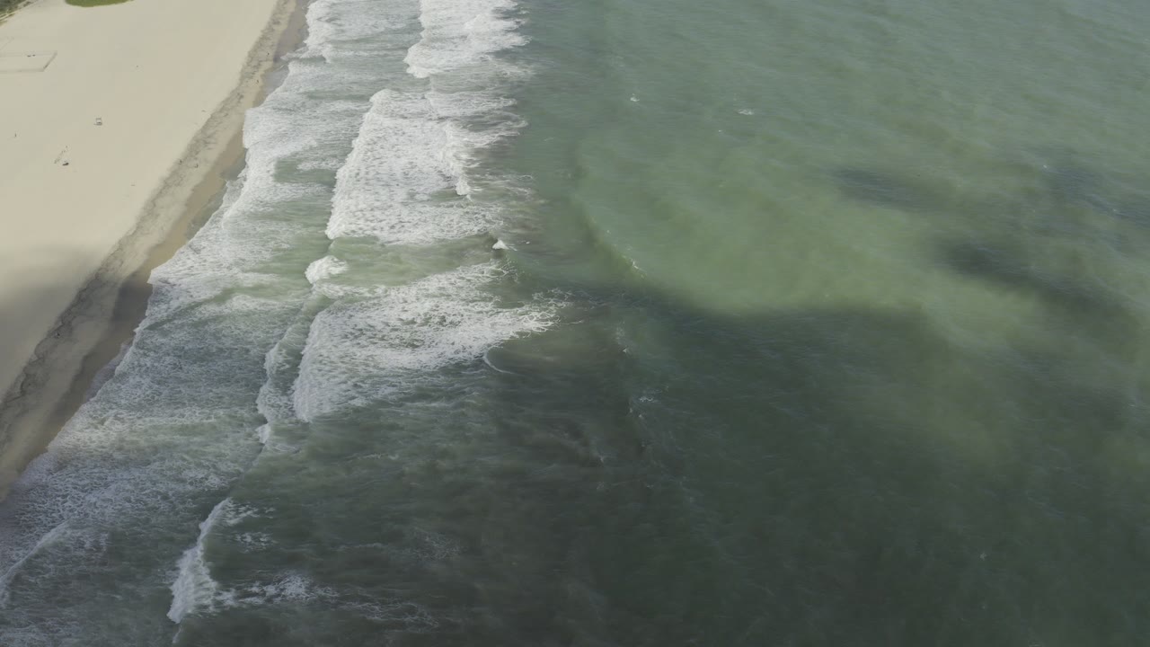 Overhead view of Los Cabos beach surf with pale sand at the left edge, green-blue water, diagonal white foam, and no visible people.