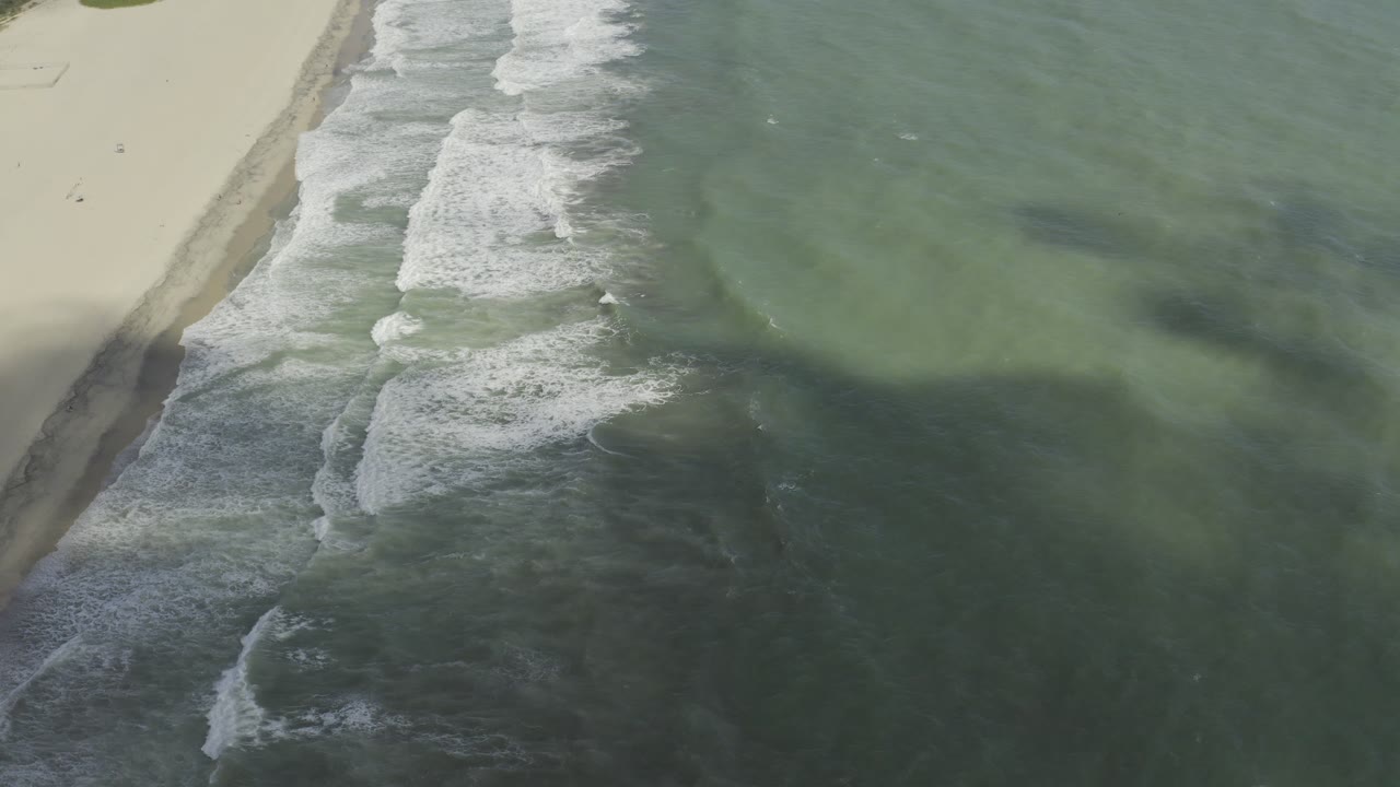 Aerial Los Cabos shoreline frame with white foam bands moving along the sand-side edge and darker green water filling most of the view, with no visible people.