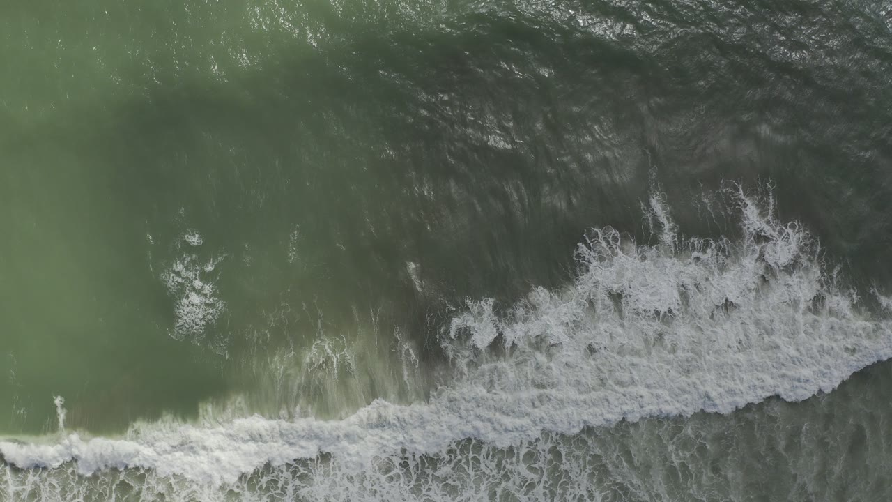 Overhead view of green ocean water in Los Cabos, Mexico, with a broad white breaker spreading across the lower edge and no visible people.