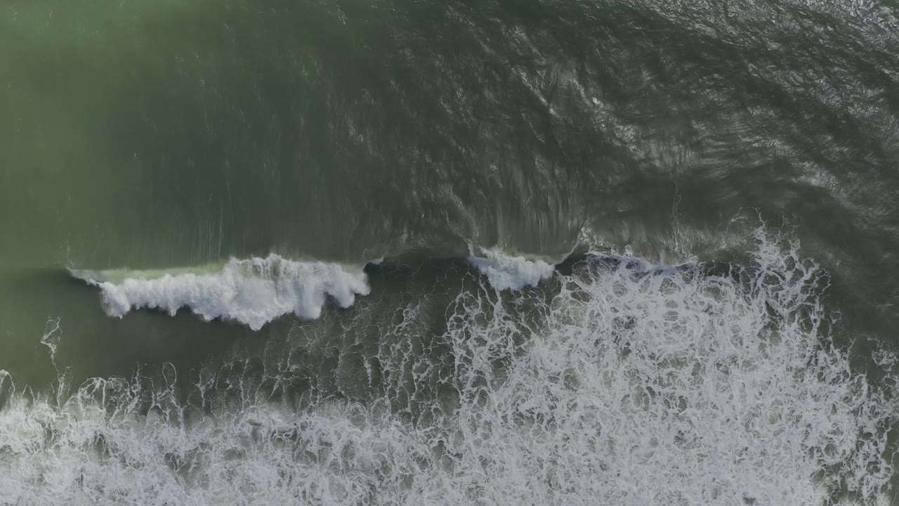 Whitewater spreads across green ocean beneath an overhead camera.