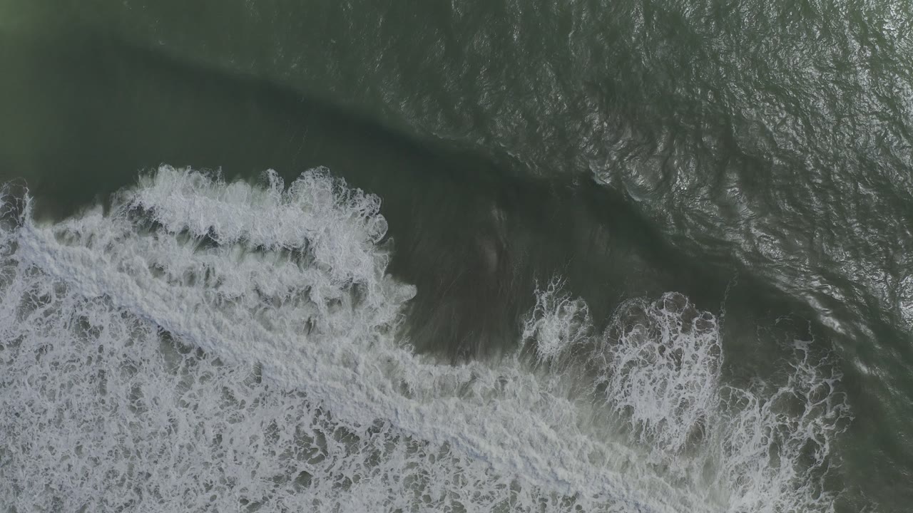 Overhead Los Cabos, Mexico view of white foam spreading diagonally across dark green ocean water, with no visible people.