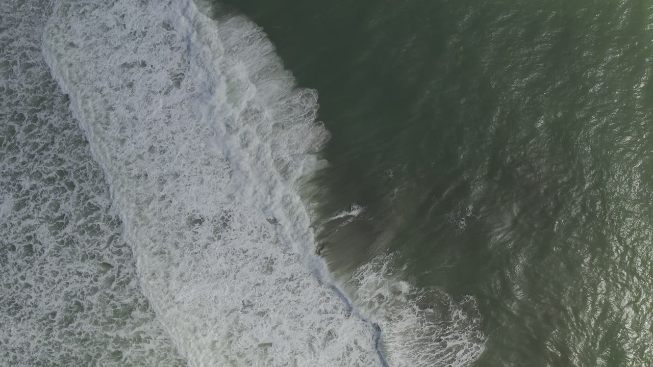 Aerial view over Los Cabos green sea with a vertical streak of breaking white foam crossing the center-left and no visible people.