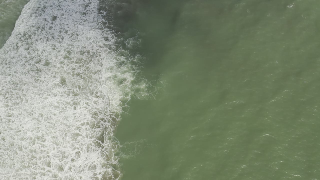 Overhead Los Cabos water view with a diagonal white foam band crossing green sea and open water to the right, no visible people.
