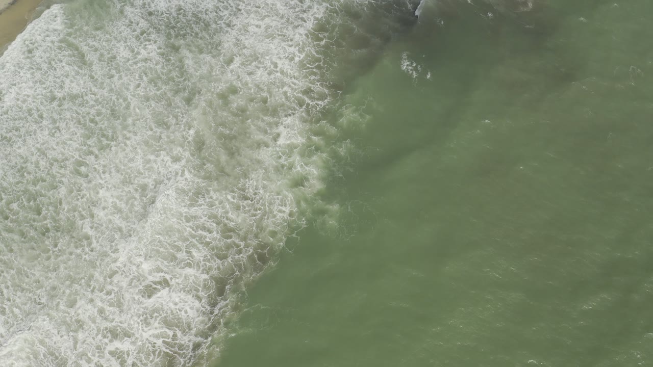 Overhead view of pale green water and white surf foam along the Los Cabos shoreline at the start of the clip, with no visible people.