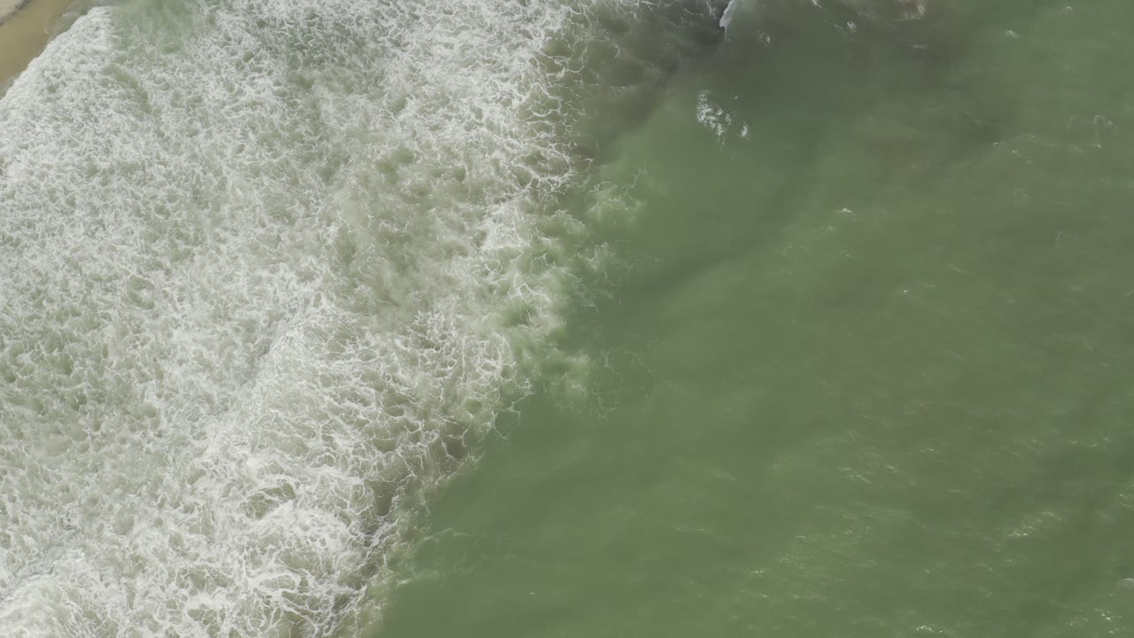 A diagonal band of white surf foam crosses pale green Los Cabos water, with the shoreline mostly outside the frame and no visible people.