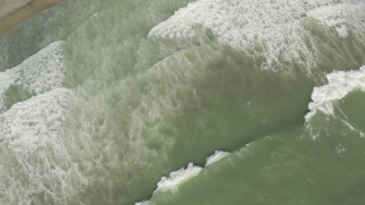 Overhead view of green ocean water and diagonal white surf near the edge of wet sand in Los Cabos, Mexico, with no visible people.
