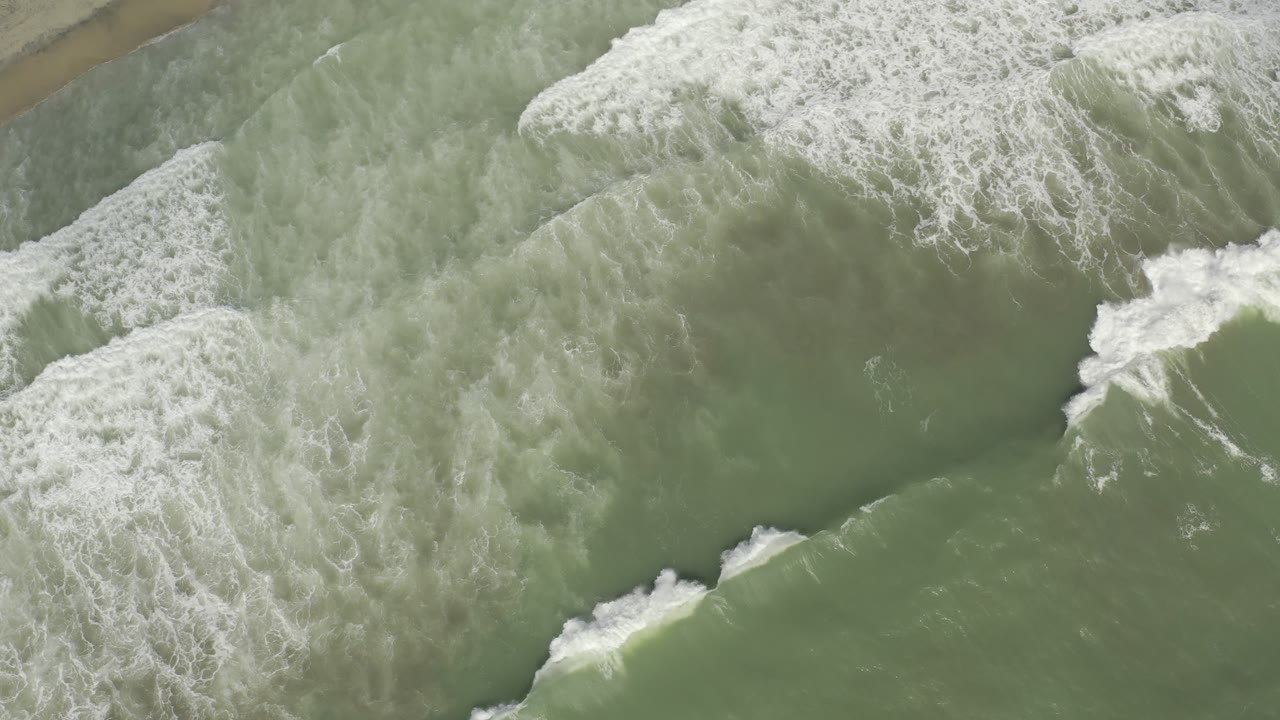 Aerial top-down frame of white foam spreading across green ocean water in Los Cabos, Mexico, with no visible people.