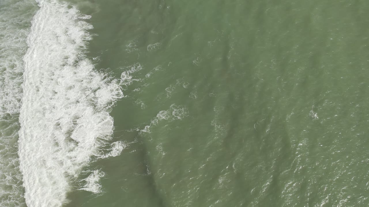 Aerial frame of white foam pushing farther into green sea from the left side in Los Cabos, Mexico, with no visible people.