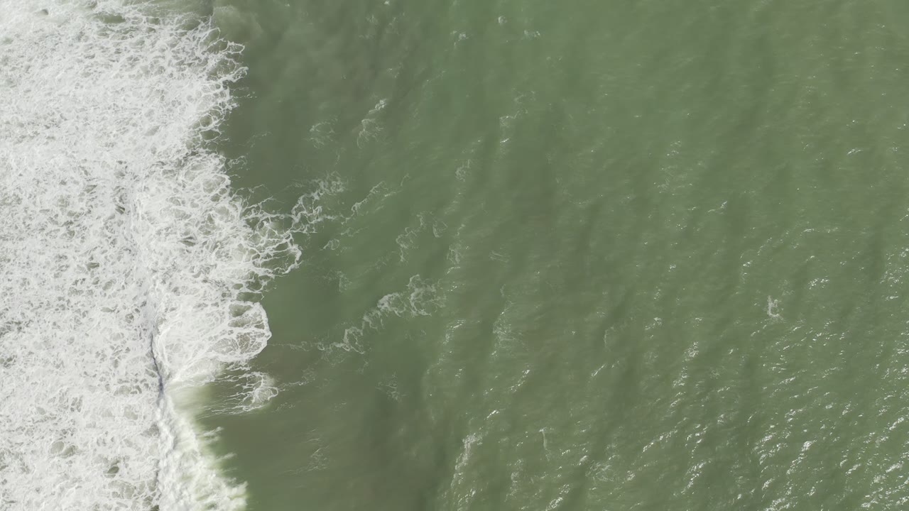 Overhead Los Cabos ocean frame with a denser white foam line pushing farther into green water and no visible people.