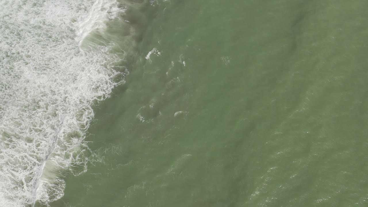 Top-down aerial view of green ocean water in Los Cabos, Mexico, with a white foam band along the left side and no visible people.