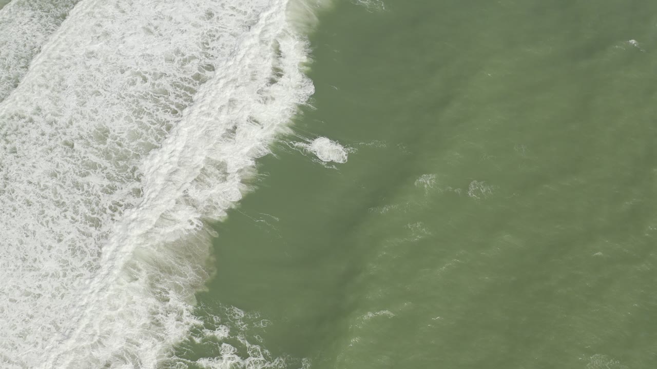 Overhead view of green-brown ocean water in Los Cabos, Mexico, with a wide white surf band along the left side and no visible people.
