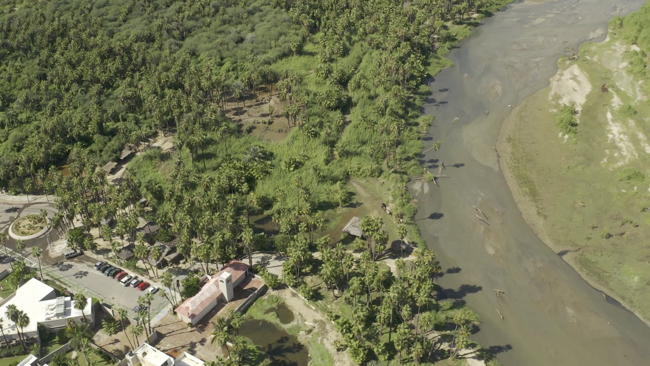 Aerial view in Los Cabos, Mexico with the river channel cutting past thick trees, roadside parked cars, low buildings, and no visible people.