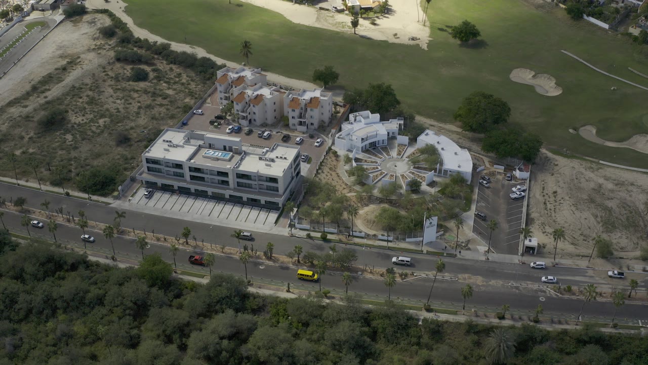 High aerial view in Los Cabos, Mexico, showing a road with cars, modern buildings, parking lots, scrubland, and a golf course beyond, with no visible people.