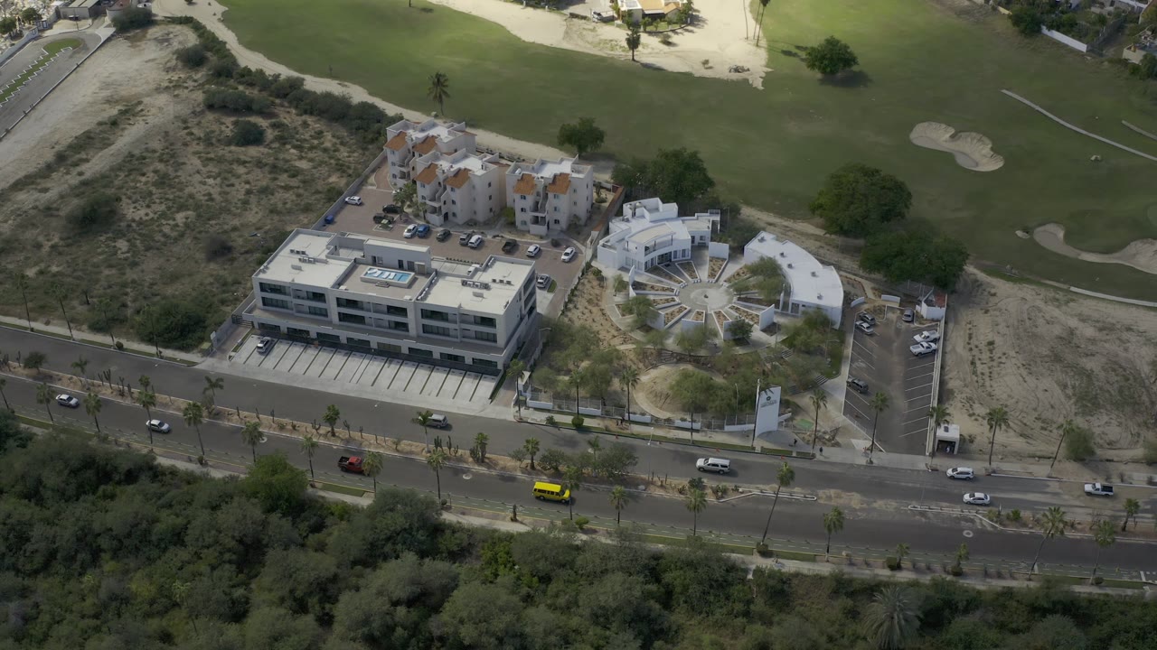 Aerial Los Cabos frame with the road running across the lower image, several cars, a flat-roofed building, parking areas, dry brush, and green golf fairways, with no visible people.