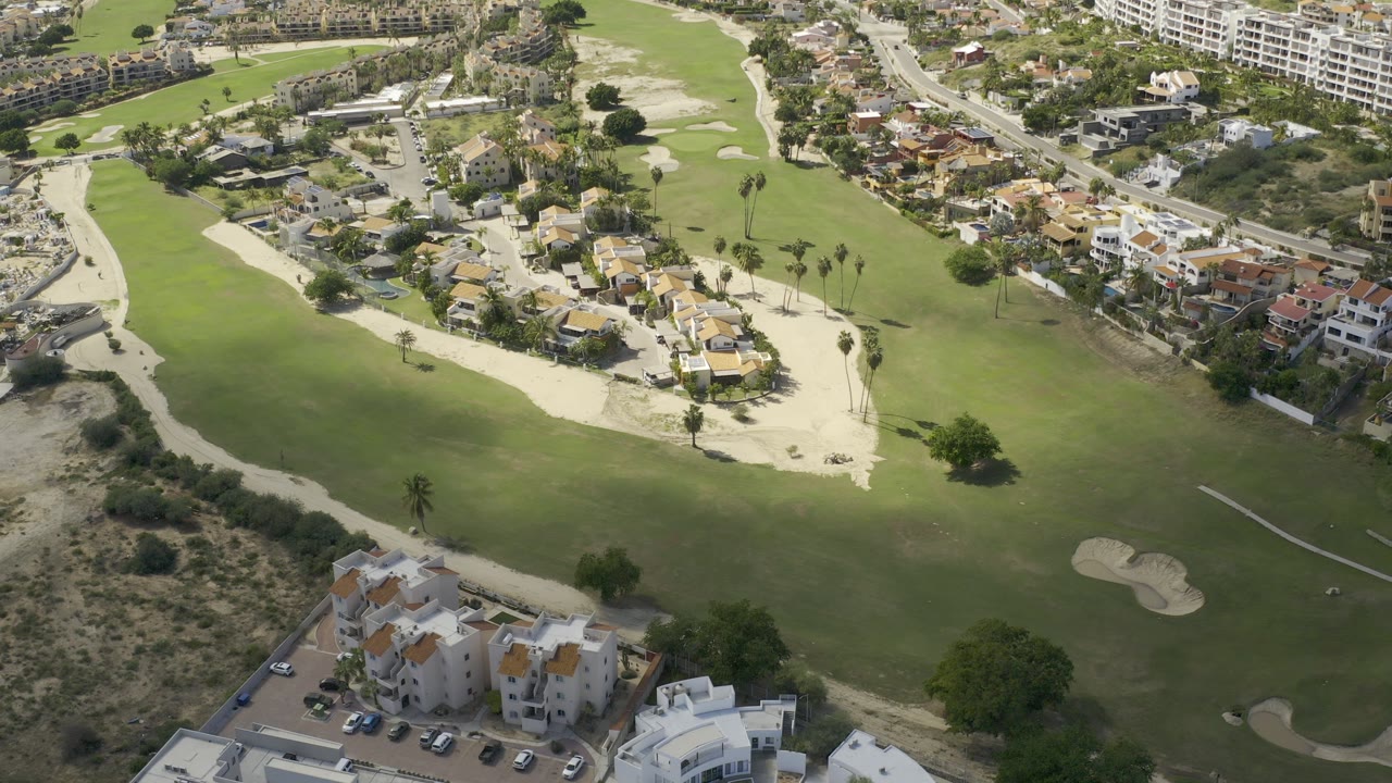 Aerial view over Los Cabos, Mexico, showing green fairways, sand bunkers, clustered homes, roadside buildings, and no visible people at the start of the clip.