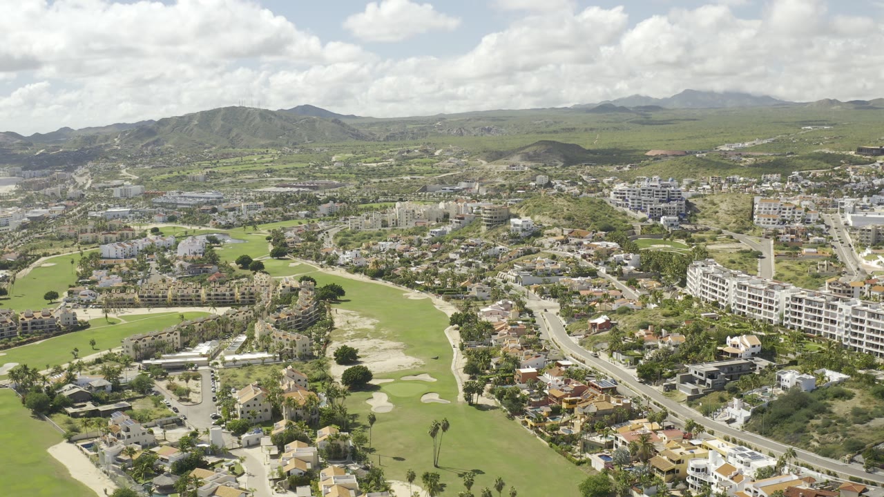 Wide aerial over Los Cabos, Mexico, showing green fairways, scattered homes, roads, mid-rise buildings, distant hills, and no visible people.