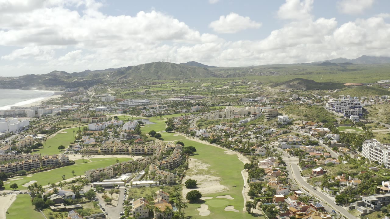 Second aerial frame in Los Cabos, Mexico, with bright fairways below, clustered homes and roads through the center, pale coastline at left, and no visible people.