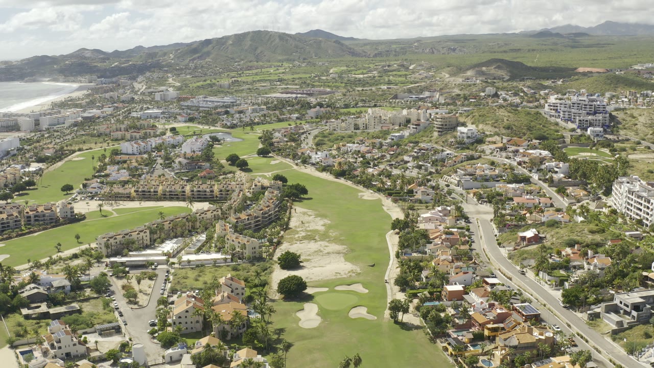 Aerial view of golf greens, houses, roads, hills, and ocean surf in the distance.