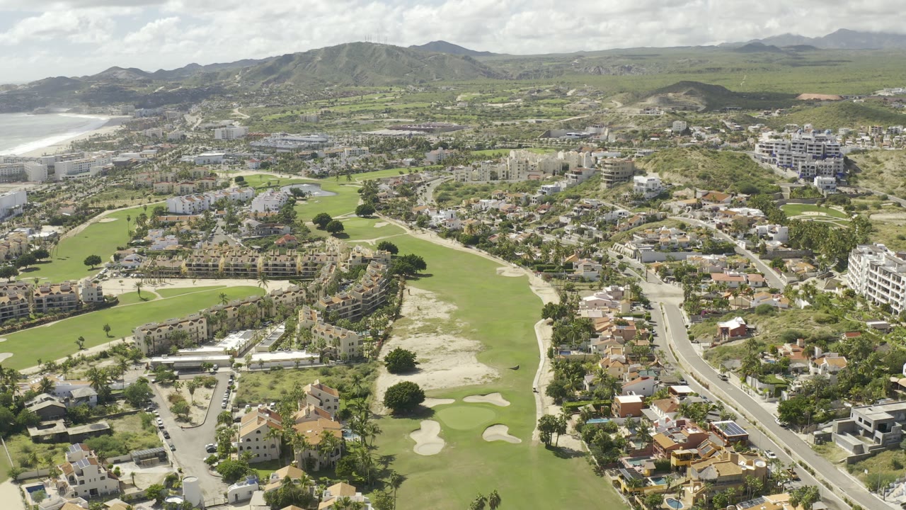 Aerial frame over Los Cabos, Mexico, showing a long green fairway beside curving residential streets, multi-story apartment blocks, open hills, distant ocean surf, and no visible people.