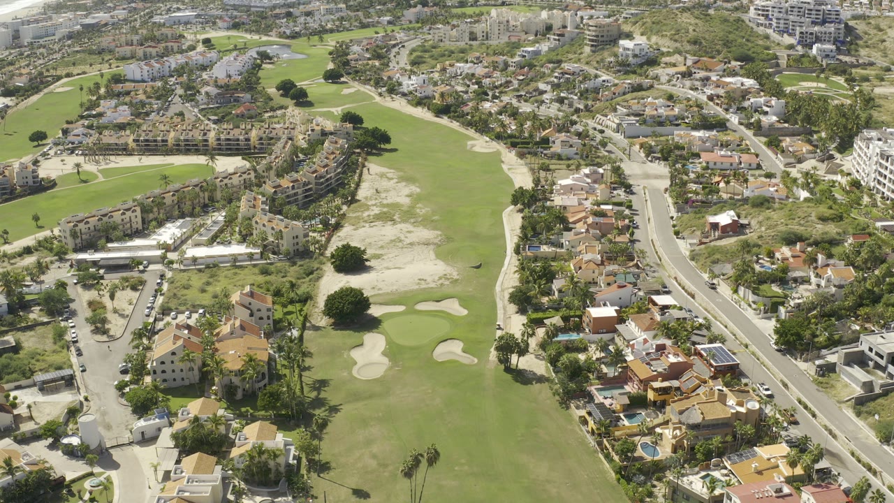 Aerial frame over Los Cabos with a green fairway and sand bunkers on the left, residential streets and tile-roof houses on the right, and no discernible faces.