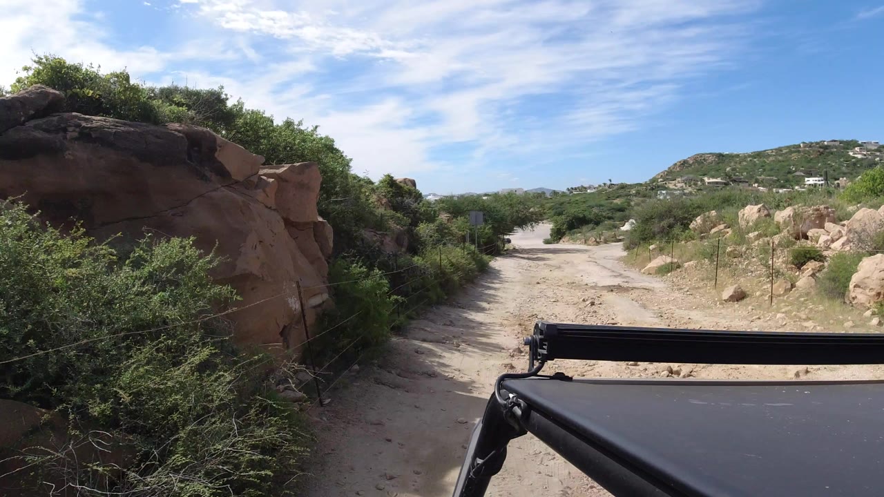 Sandy road beside rocks and green shrubs under blue sky.