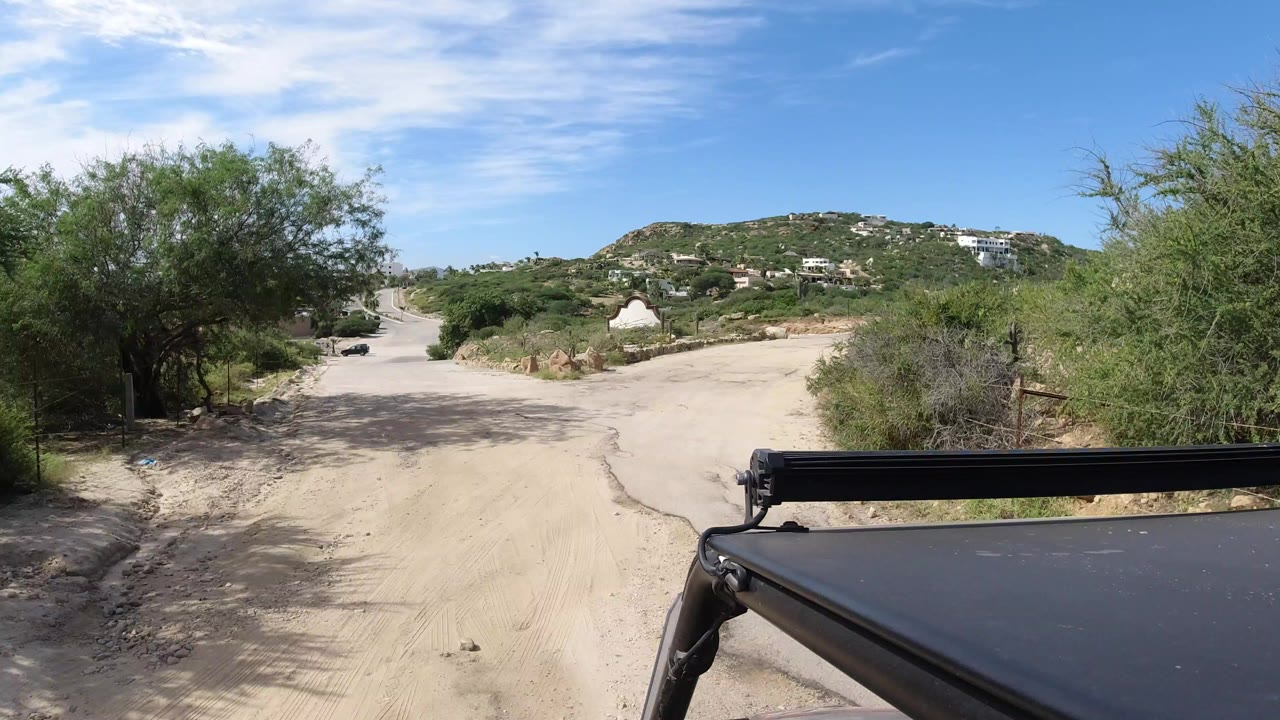Sandy road seen past a black vehicle roof and desert shrubs.