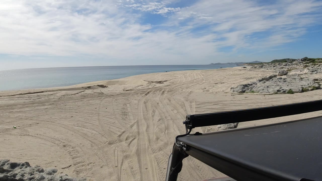 Los Cabos sand track continues along the shoreline, with the Jeep roof edge low in frame, the sea stretching left, dunes and scrub on the right, and no visible people.