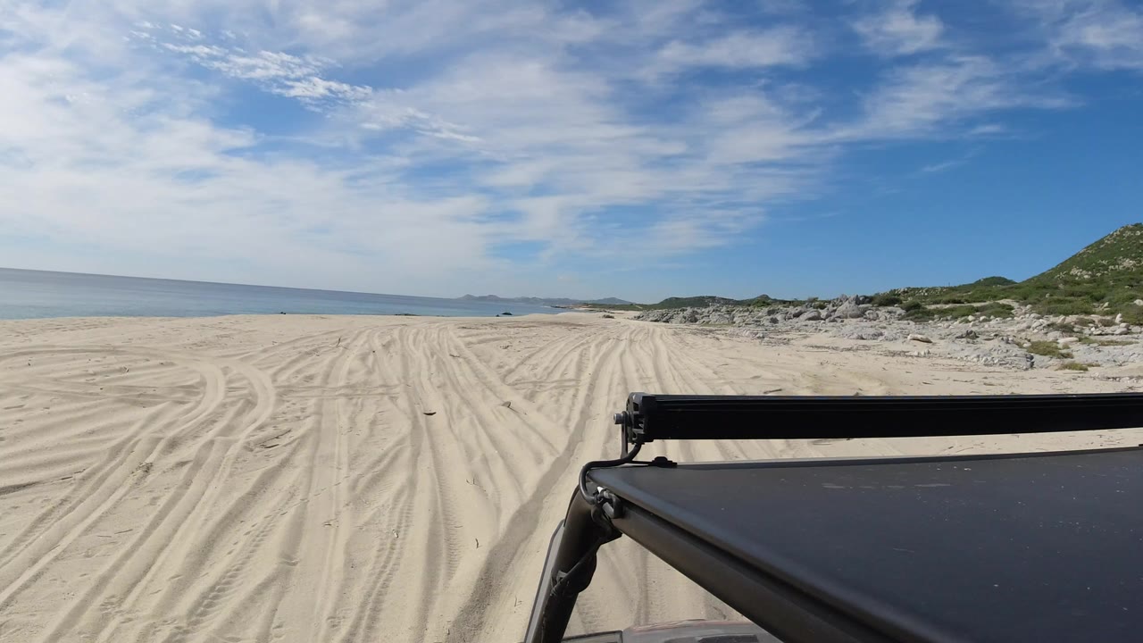 Vehicle foreground on a sandy beach with ocean and cloudy blue sky.
