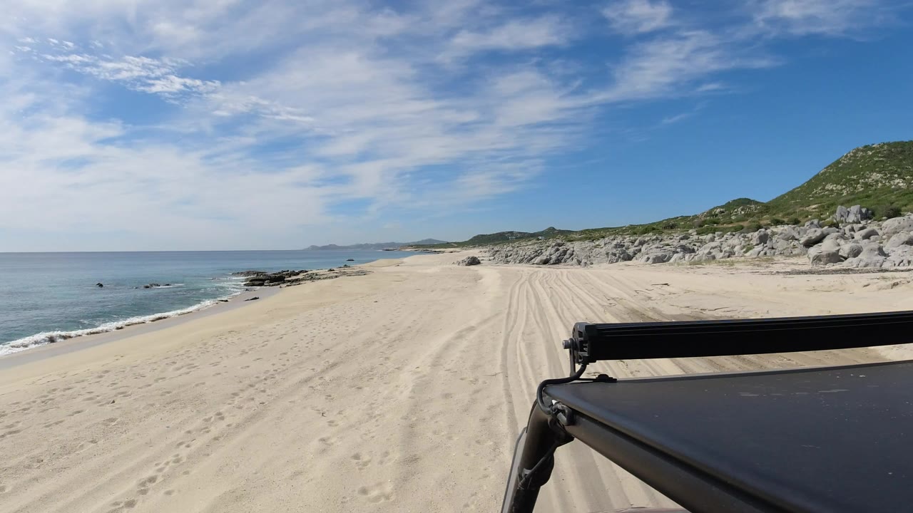 Beach track with ocean, rocks, green hill, and vehicle roof edge.