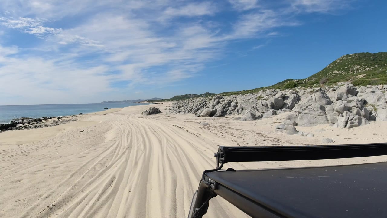 Vehicle foreground facing sandy beach tracks beside ocean and rocks
