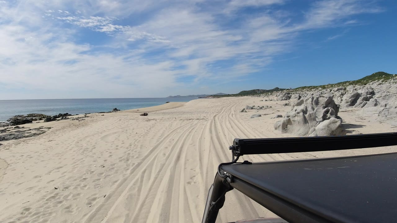 Black SUV hood and light bar frame a sandy Los Cabos beach track with ocean on the left, white boulders and dunes on the right, and no visible people.