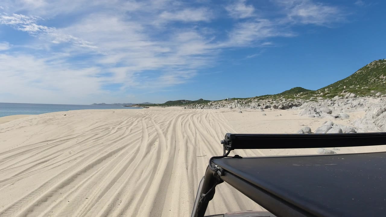 One second into the Los Cabos clip, the truck edge remains low in frame while parallel sand tracks lead toward distant coastline, with sea to the left and no visible people.