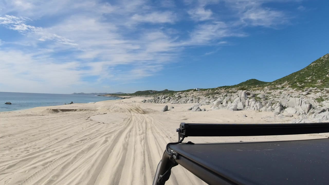 Opening Los Cabos frame with rutted pale sand ahead, calm ocean on the left, rocky dune hills on the right, a black vehicle edge in the foreground, and no visible people.