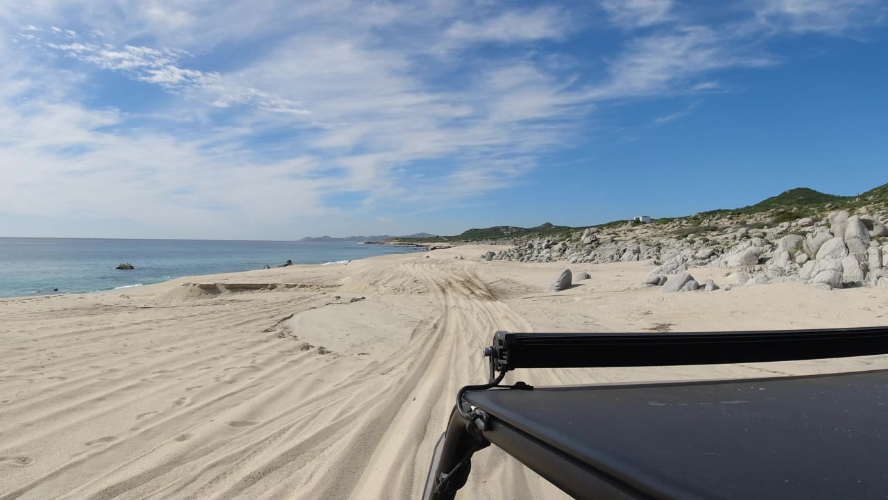 Wide coastal sand path with vehicle light bar, ocean, rocks, and low hills.