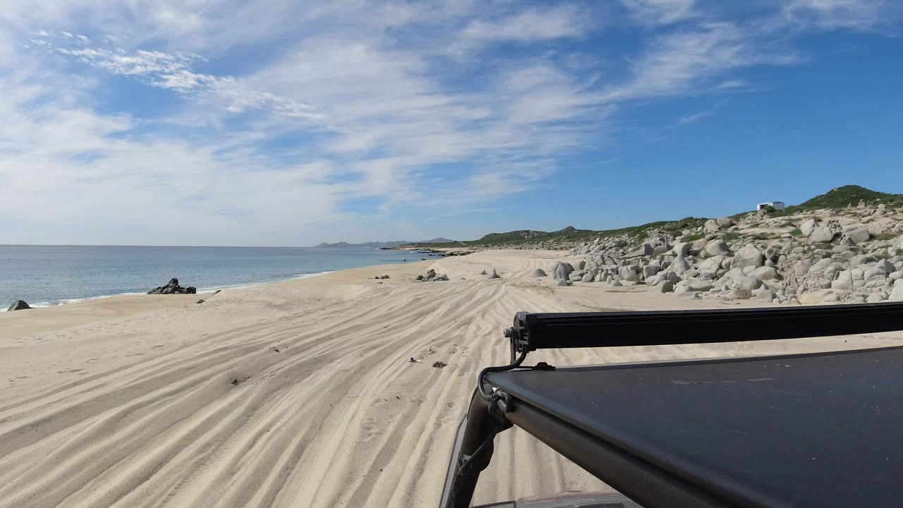 Start of a Los Cabos beach driving view with vehicle hood and light bar in the foreground, tire tracks in pale sand, blue sea at left, rocky shoreline at right, and no visible people.