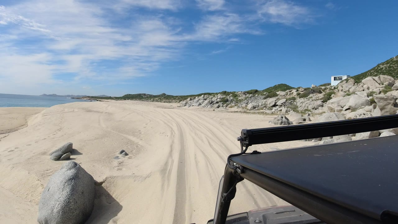 At 0:01 in Los Cabos, Mexico, the vehicle edge remains low in frame as the sandy track curves between the waterline and rocky dunes, with no visible people.