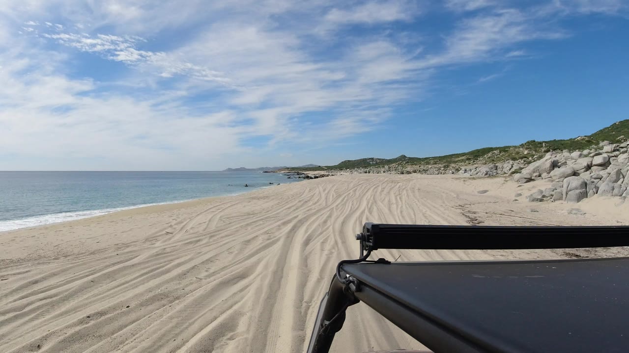 Opening Los Cabos beach-drive frame with pale tire-marked sand ahead, blue ocean on the left, rocky dunes on the right, a dark foreground rig edge, and no visible people.