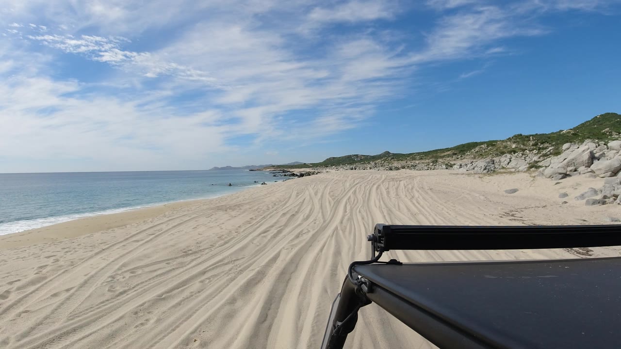 Sandy beach tracks beside clear ocean and low coastal hills.