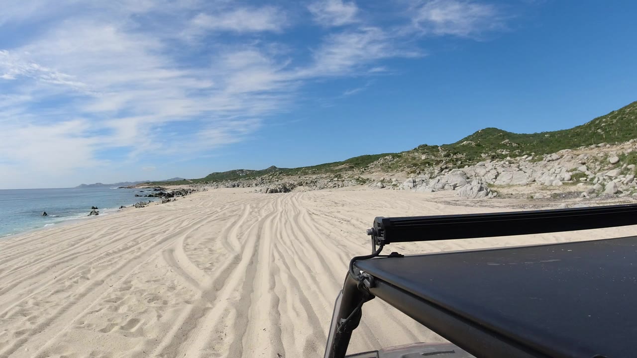 Opening view from a vehicle on a Los Cabos sandy beach, with ocean to the left, tire tracks ahead, rocky dunes to the right, and no visible people.