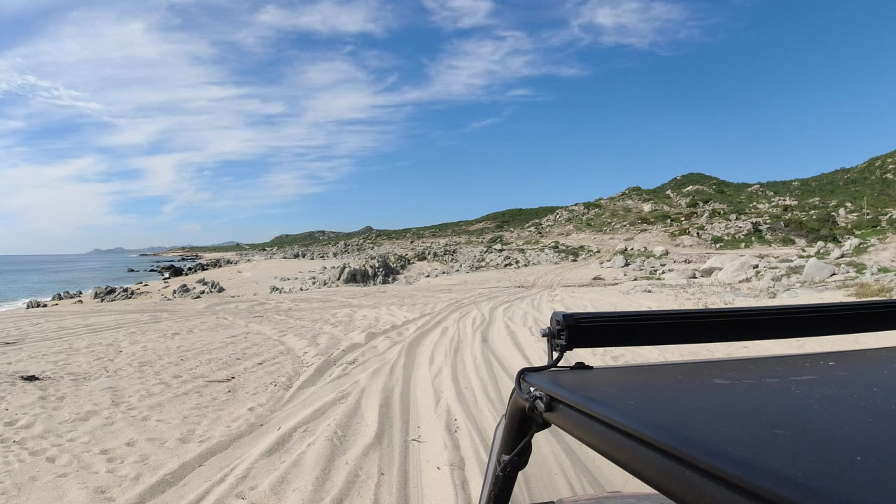 Black SUV foreground on sandy coastal road with rocks and low hills.