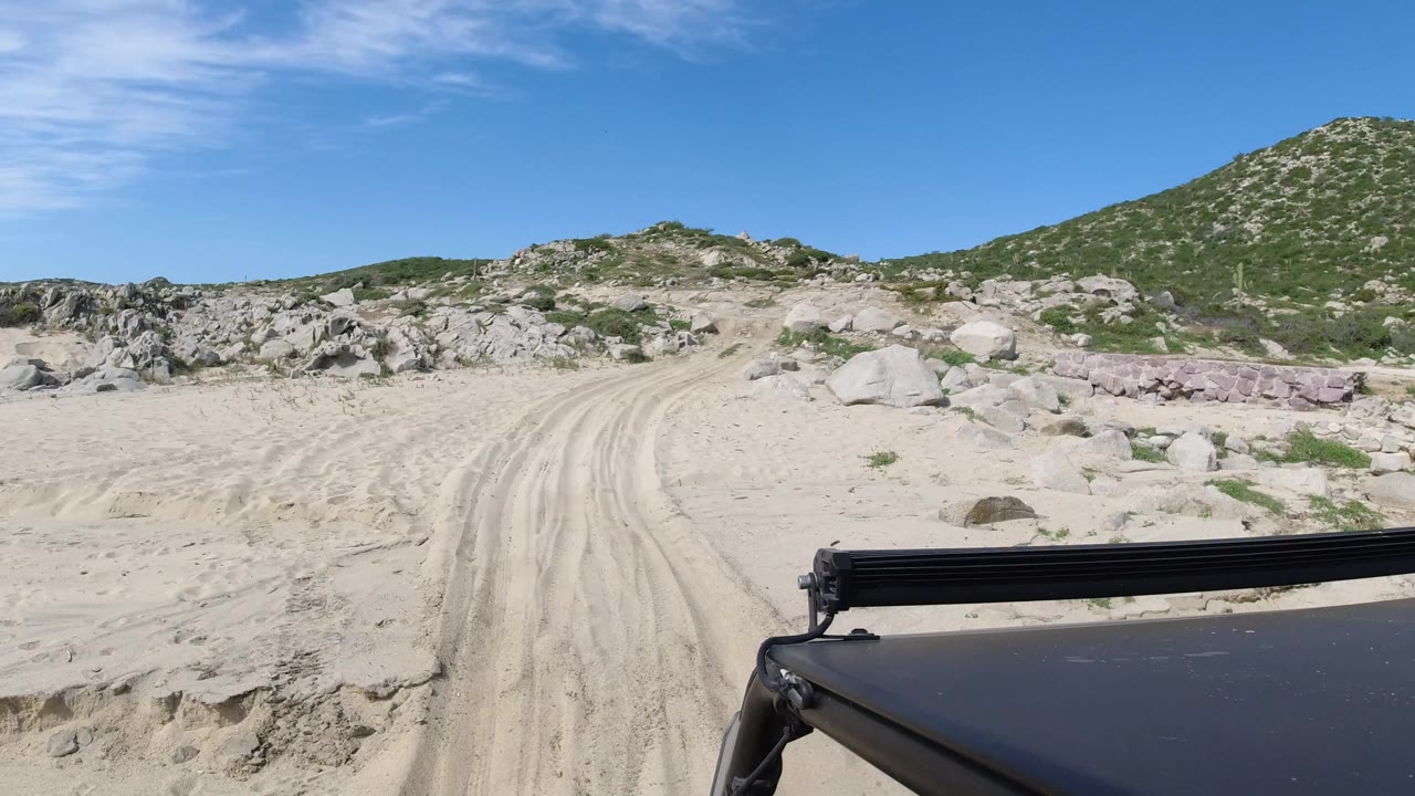 A rutted sand path leads between boulders and scrub in Los Cabos, Mexico, while the lower frame includes the vehicle roof and no visible people.