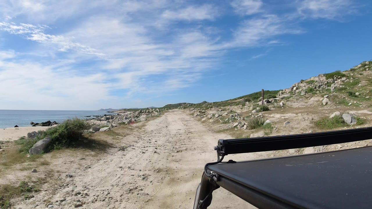 Sandy coastal dirt road beside ocean and rocks with car roof in foreground.