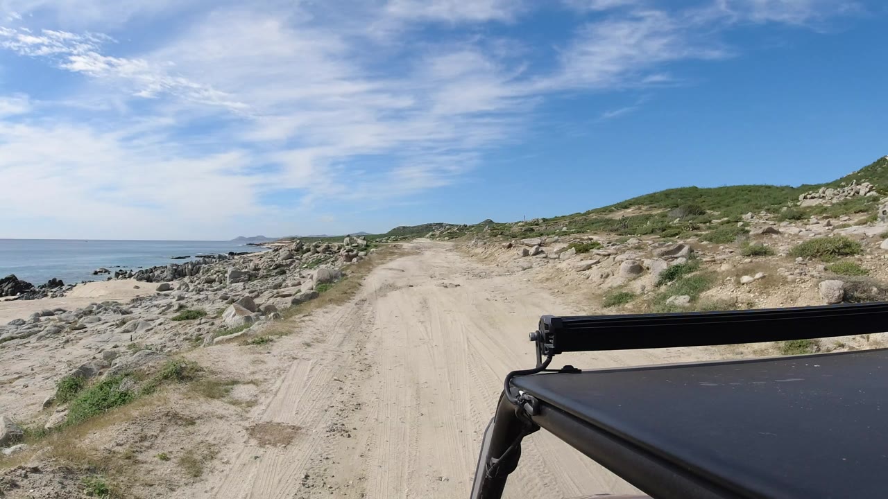 Opening view of a sandy road along the Los Cabos shoreline, with blue water to the left, rocky ground and low green brush ahead, and no visible people.