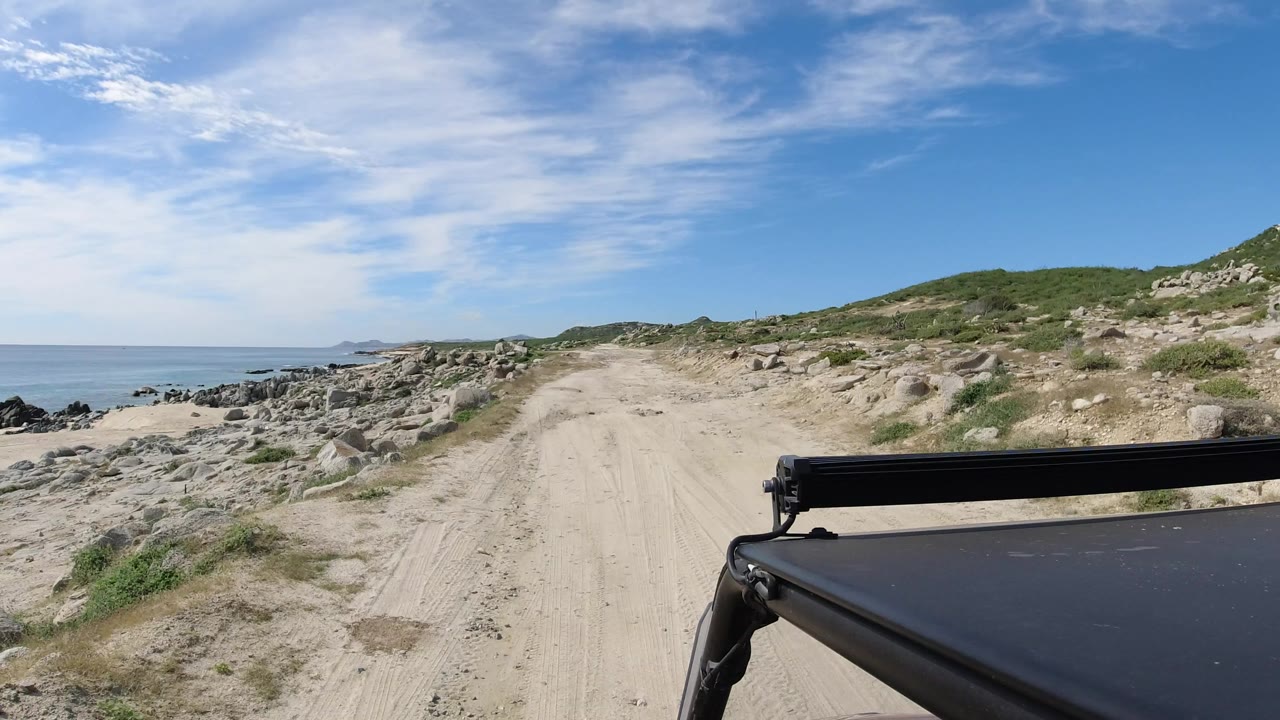 The Los Cabos dirt track continues forward beside scattered shoreline rocks, with the ocean visible on the left and the black vehicle edge fixed at lower right, no visible people.