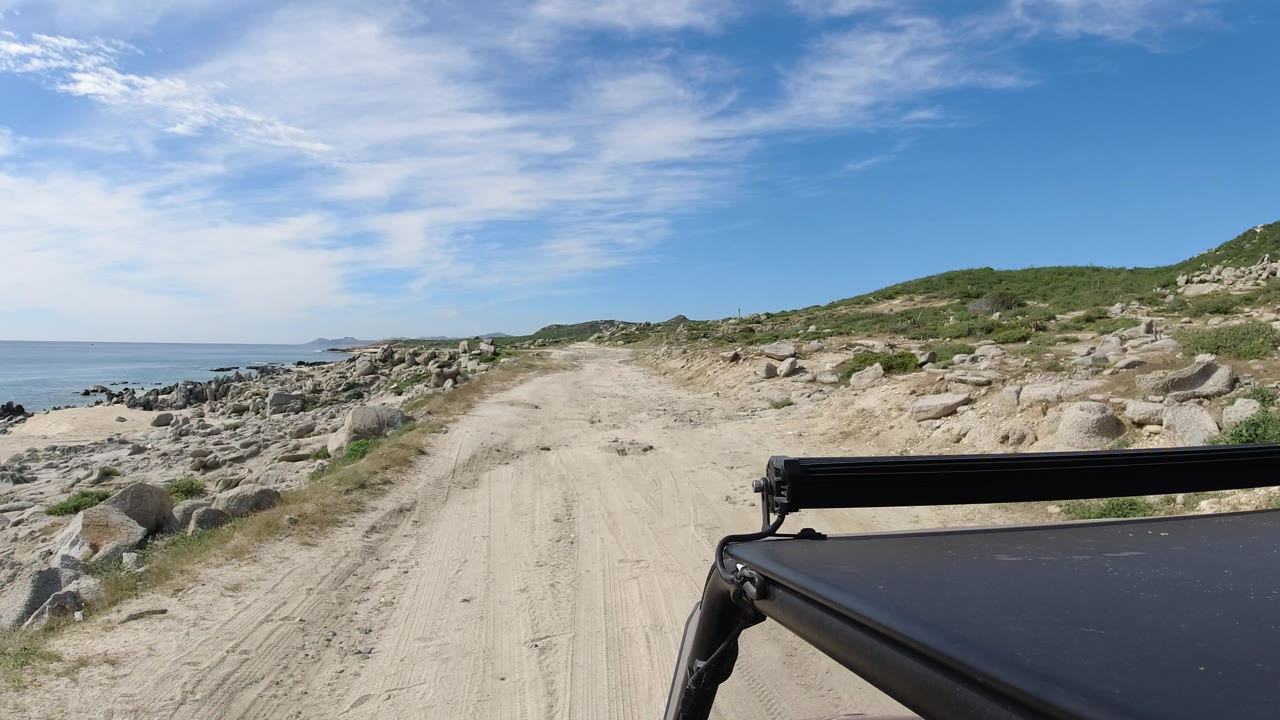 Vehicle-side view of a sandy track between ocean rocks and scrubby hillside.