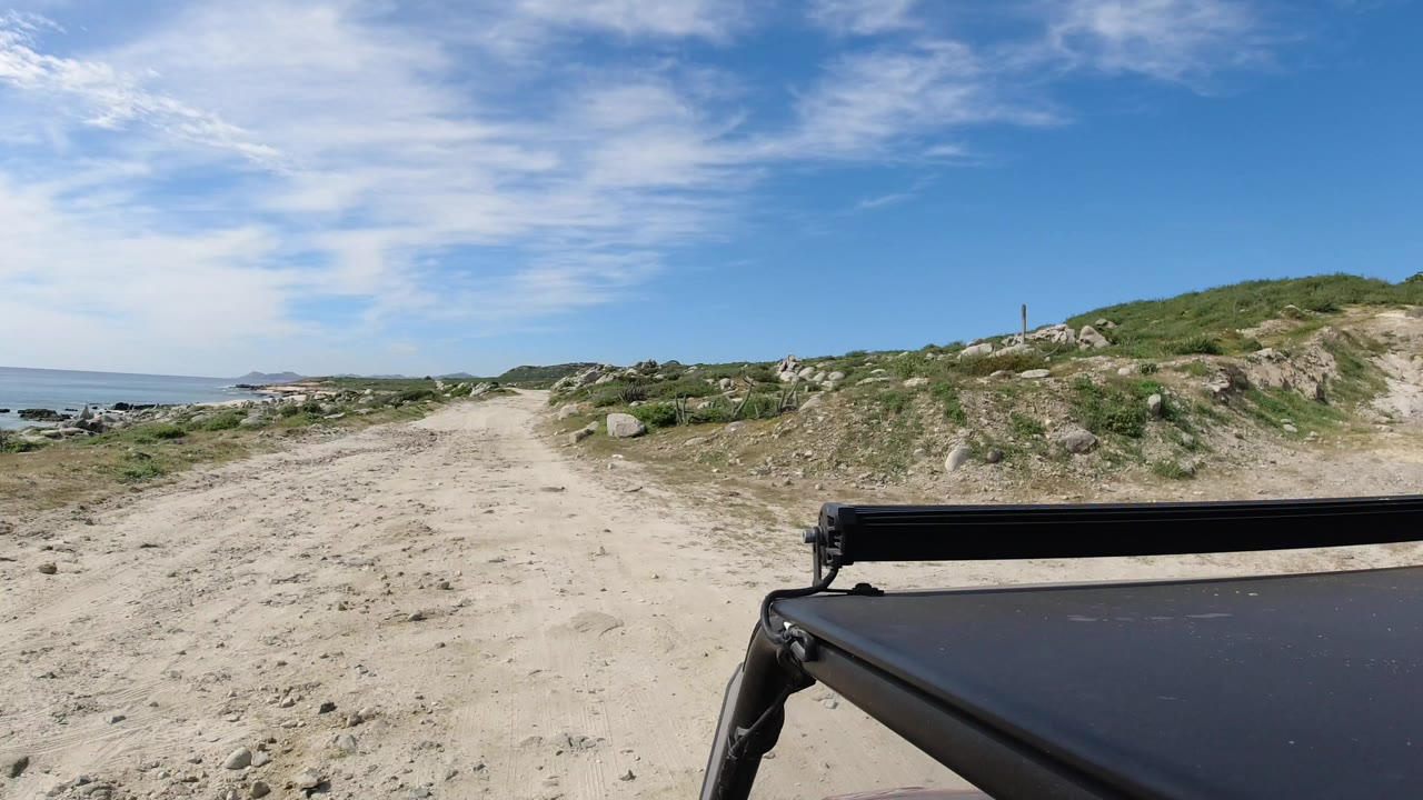 Sandy coastal road with ocean to the left and vehicle roof in foreground